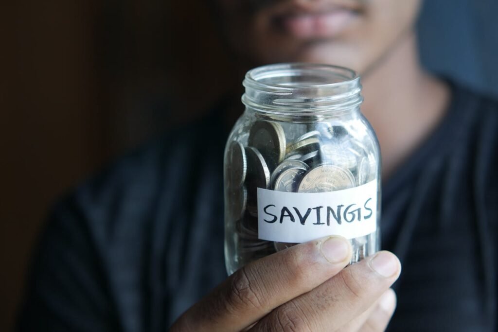 A close-up image of a person's hand holding a jar full of coins labeled 'Savings'.